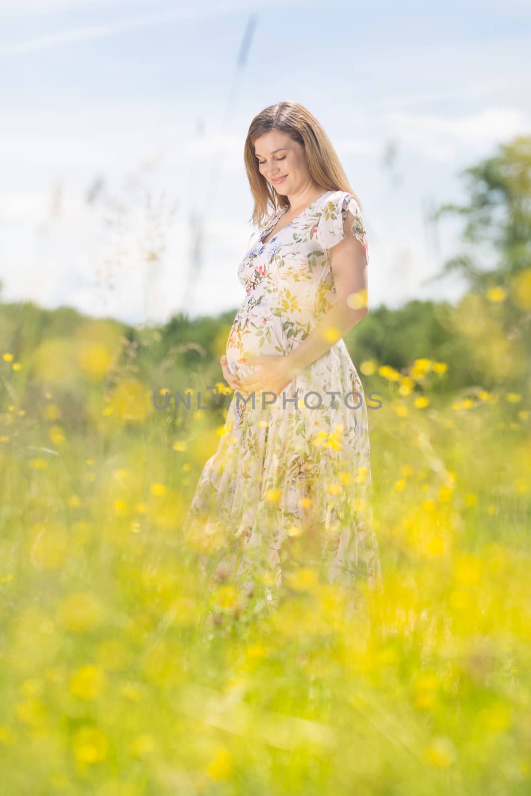 Portrait of beautiful happy pregnant woman smiling in white summer dress relaxing in meadow full of yellow blooming flowers. Concept of healthy maternity care.