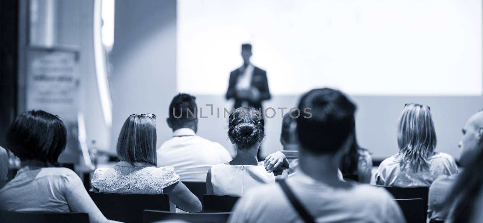 Speaker giving talk at business event. Audience at the conference hall. Business and Entrepreneurship concept. Focus on unrecognizable people in audience. Blue toned grayscale image.