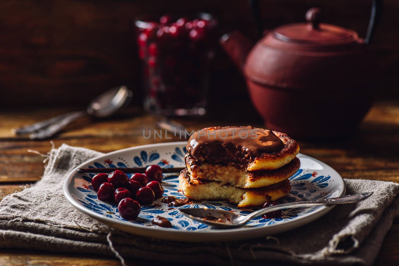 Quark Pancakes with Chocolate Topping, Frozen Cherry and Vanilla Pod. Tea Pot with Spoons and Glass of Berries on Background.