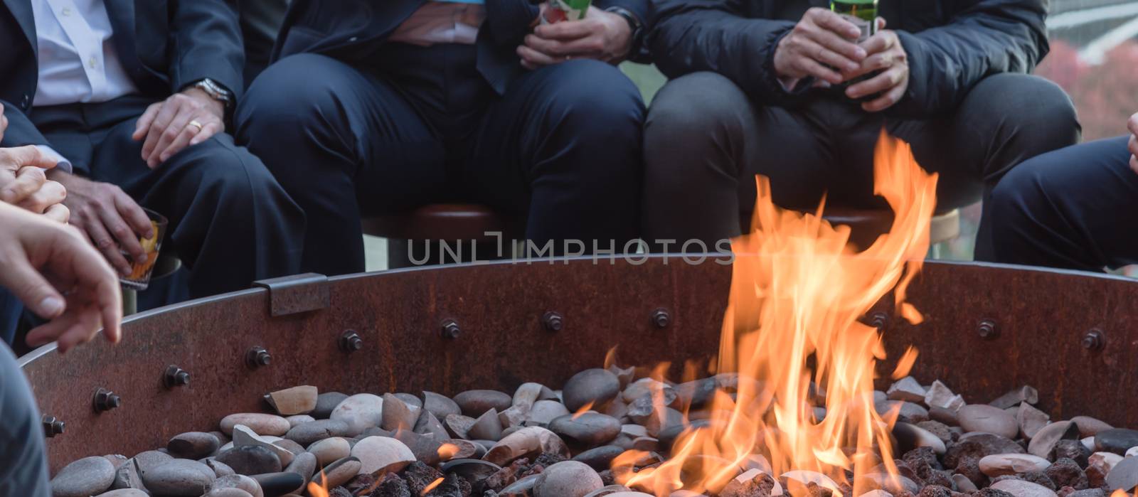 Panorama view group of Caucasian businessmen in formal dress gathering over the round fire pit after work at upscale bar in Chicago. Business people watches, leather shoes drinking beer, wine