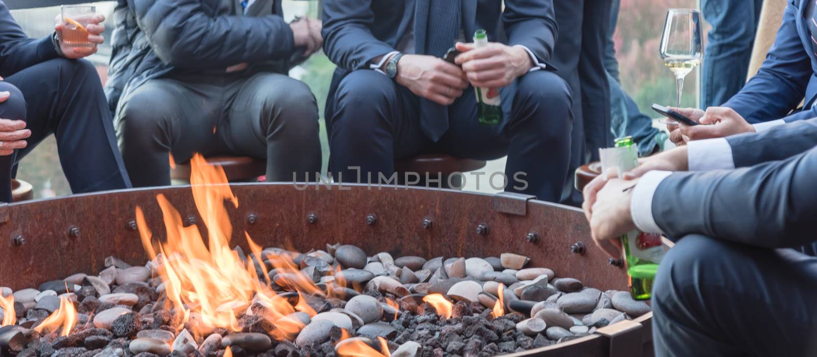 Panorama view group of Caucasian businessmen in formal dress gathering over the round fire pit after work at upscale bar in Chicago. Business people watches, leather shoes drinking beer, wine