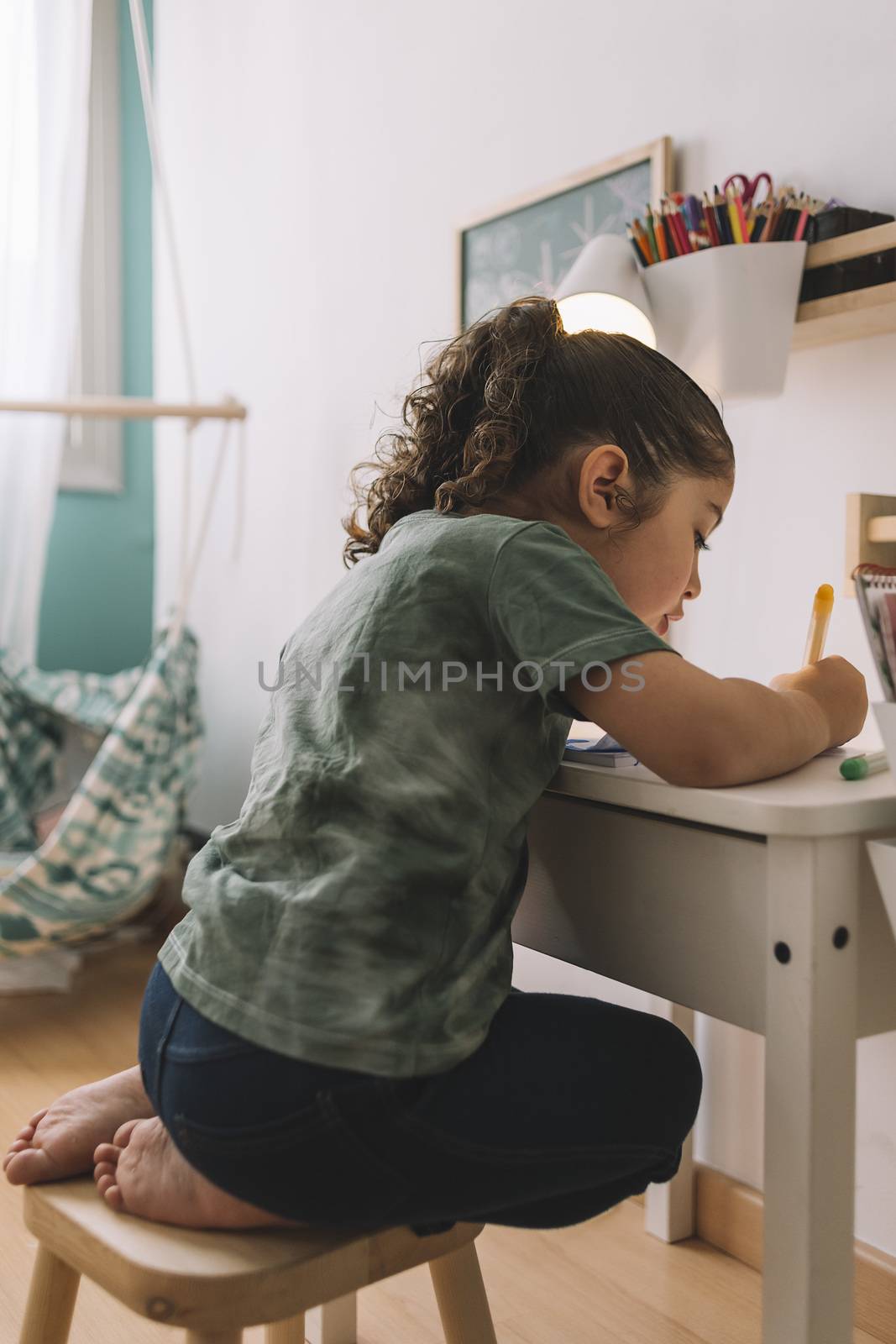 vertical photo of a little girl that draws concentrated at home with color markers, she is drawing on the desk in her room