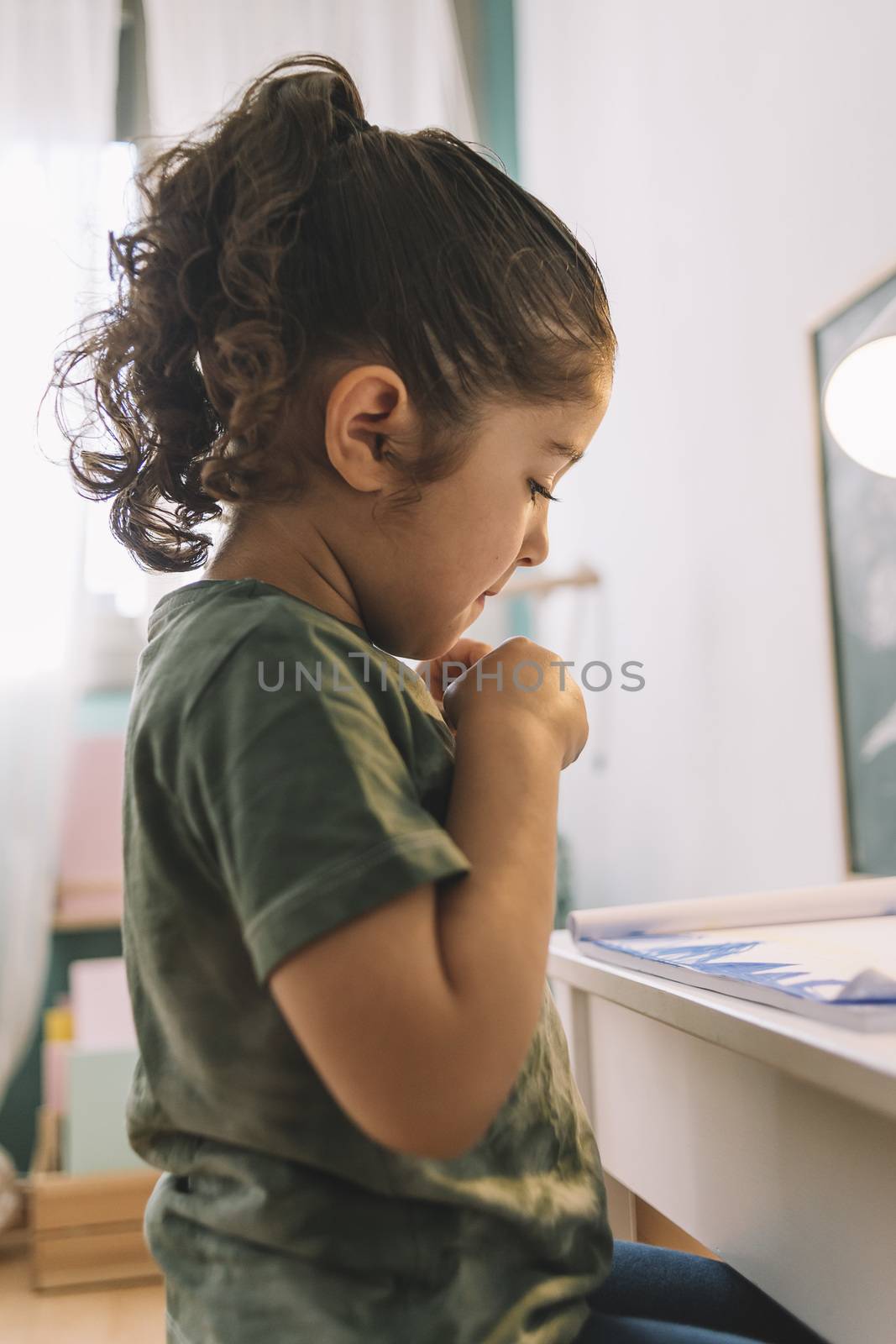 vertical photo of a little girl looking at the draw she just made at home with color markers, the draw is on the desk in her room