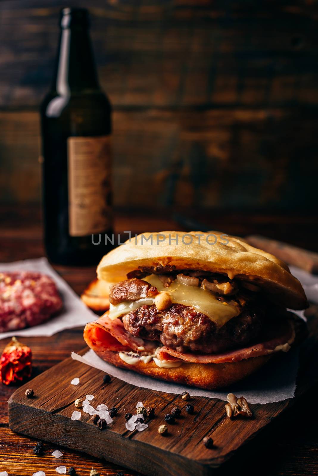 The Casanova Burger on Cutting Board with Beef Patty, Wisconsin Swiss Cheese, Ham, Sauteed Mushrooms, Dijon Mustard, Mayonnaise and Potato Roll. Bottle of Craft Beer on Background.