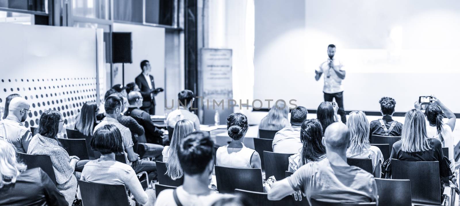 Male speaker giving a talk in conference hall at business event. Audience at the conference hall. Business and Entrepreneurship concept. Focus on unrecognizable people in audience. Blue toned.