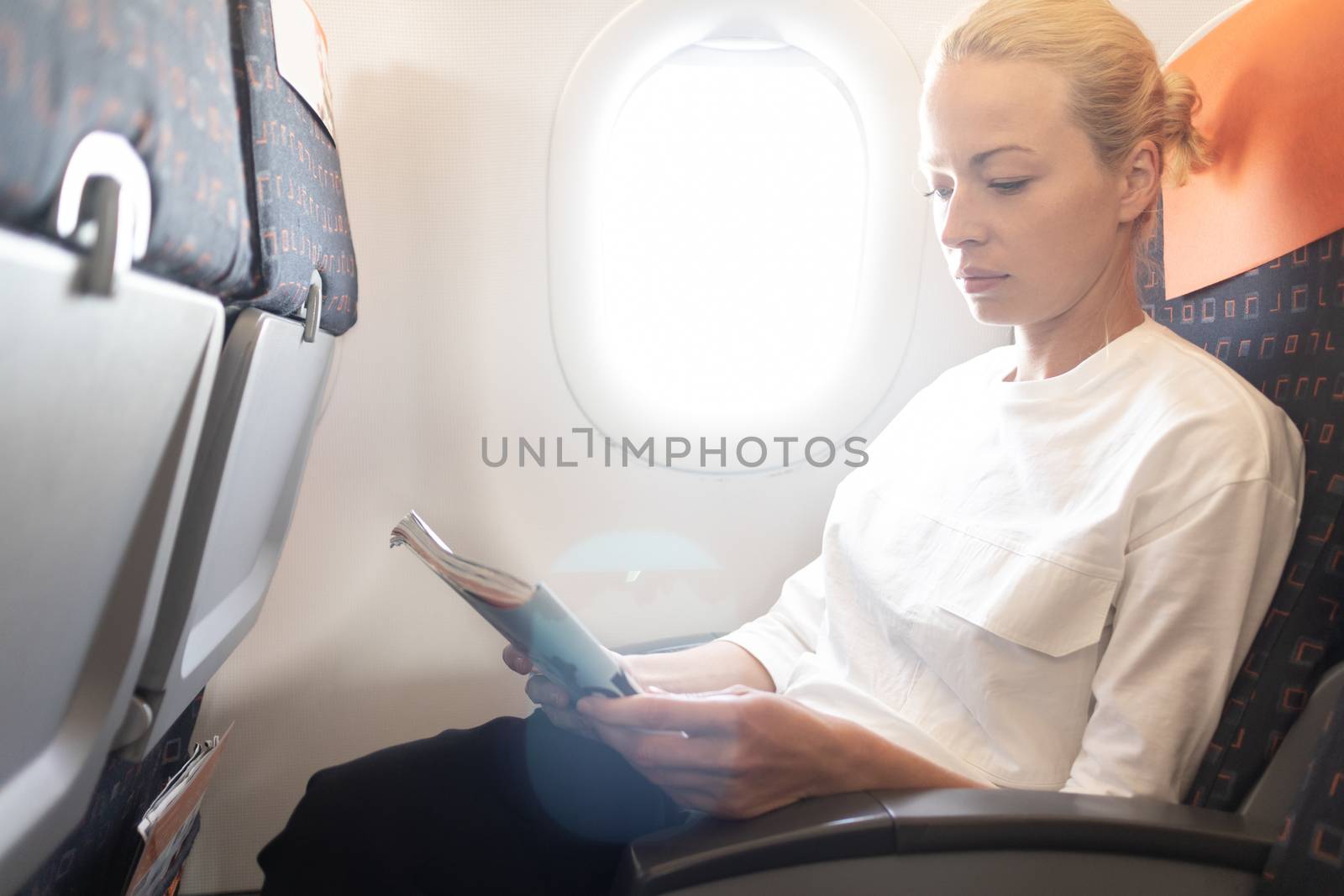 Woman reading in flight magazine on airplane. Female traveler reading seated in passanger cabin. Sun shining trough airplane window.