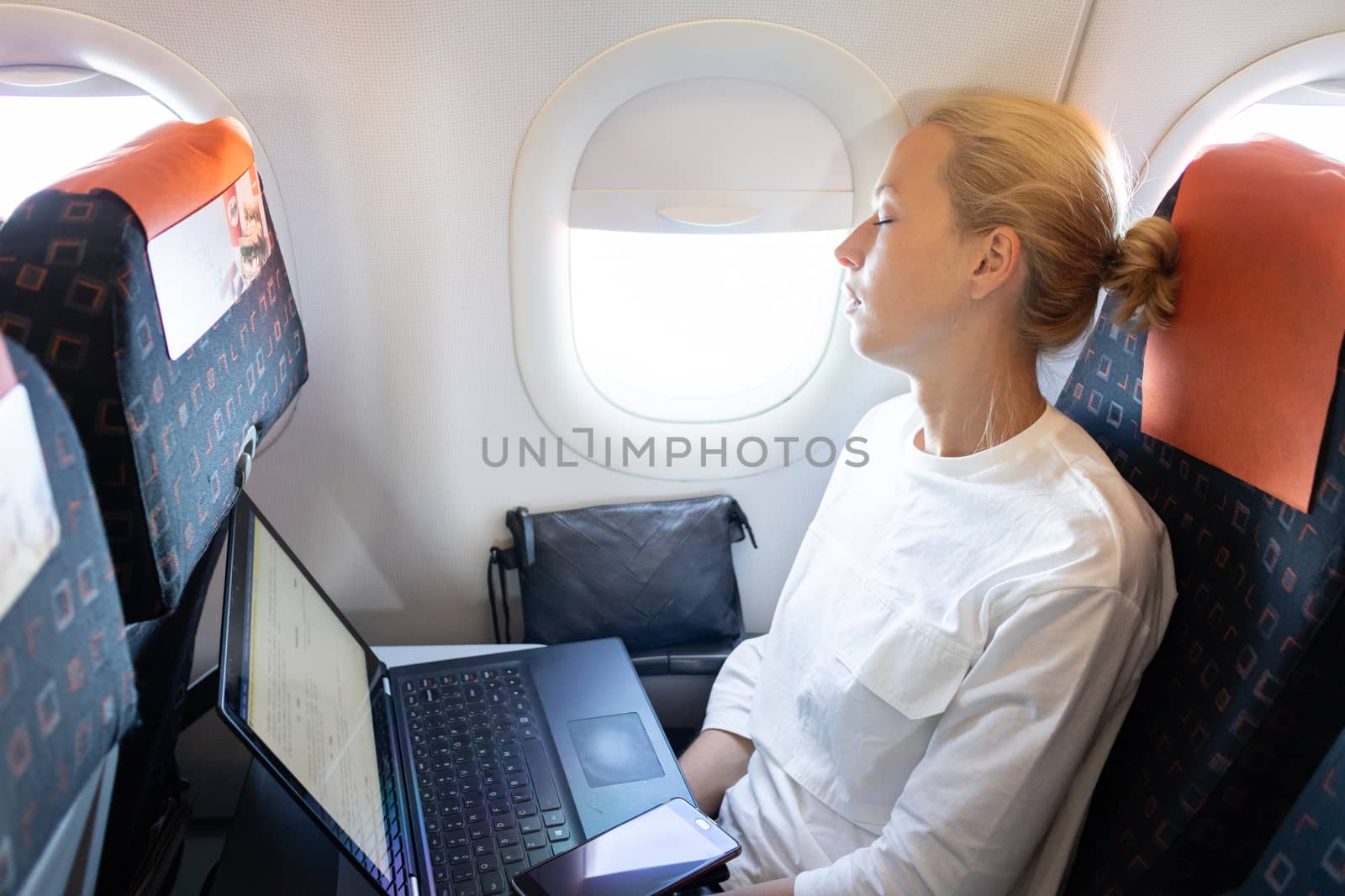 Tired business woman napping on airplane during her business trip woking tasks. Female passenger in flying aircraft sleeps next to the window.