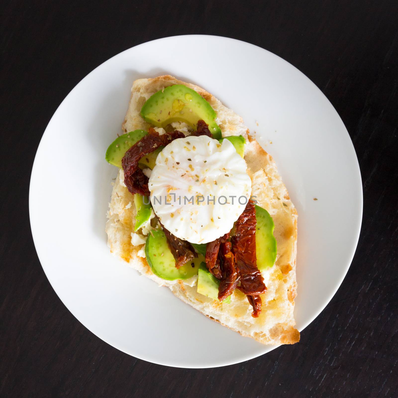 Poached egg on a toasted bun with avocados and dried tomatoes on white plate.