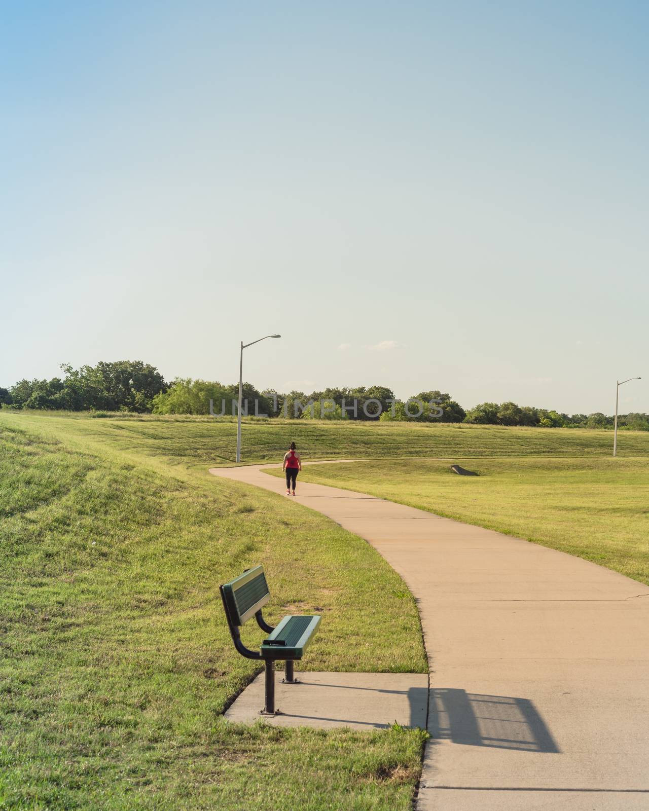 Curved pathway with bench in a hillside urban park near Dallas, Texas, America. Rearview of chubby lady jogging with headphone near lamppost.
