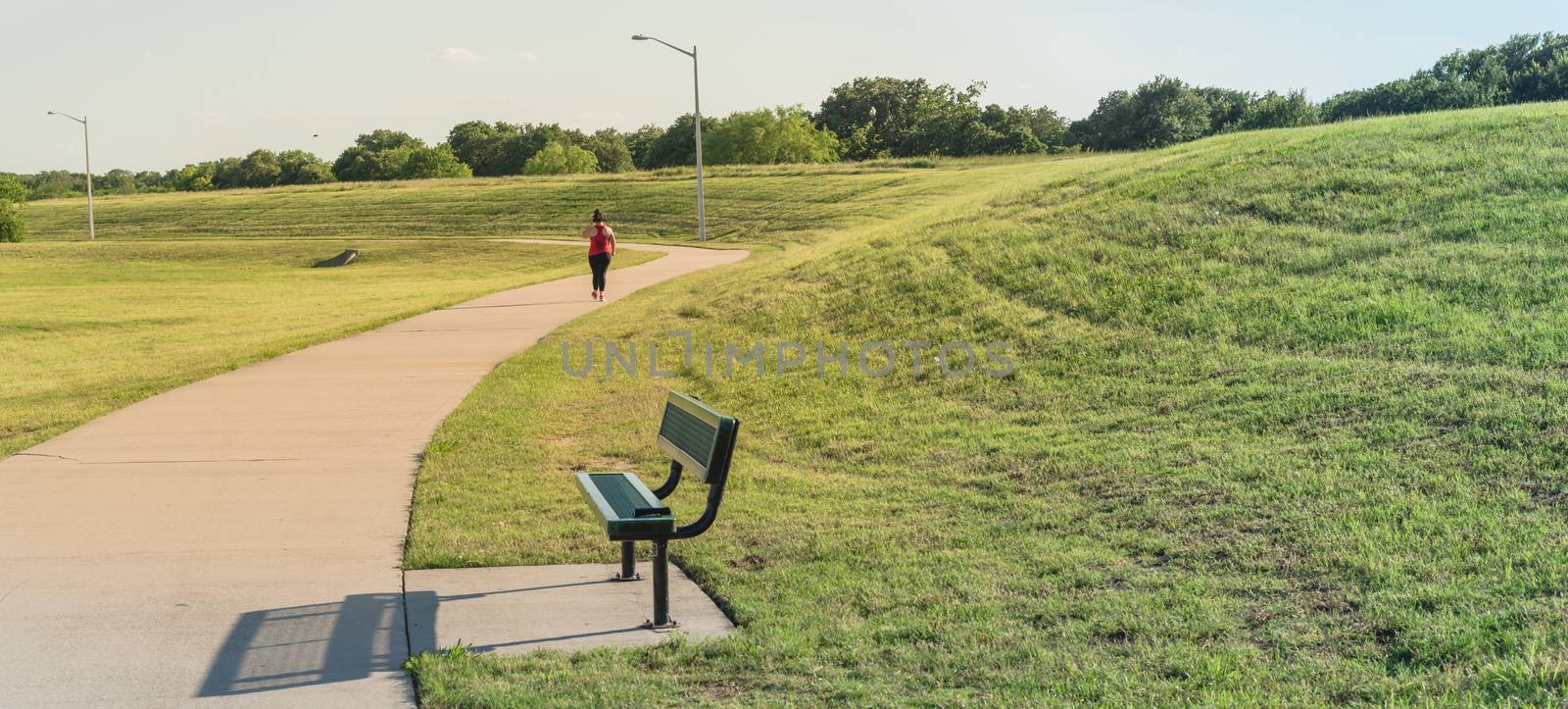 Curved pathway with bench in a hillside urban park near Dallas, Texas, America. Rearview of chubby lady jogging with headphone near lamppost.