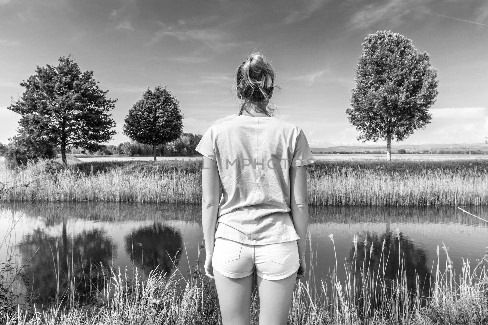 Young woman wearing casual summer clothes enjoying beautiful view of peaceful countryside. Trees on the meadow reflecting in the river. Black and white image.