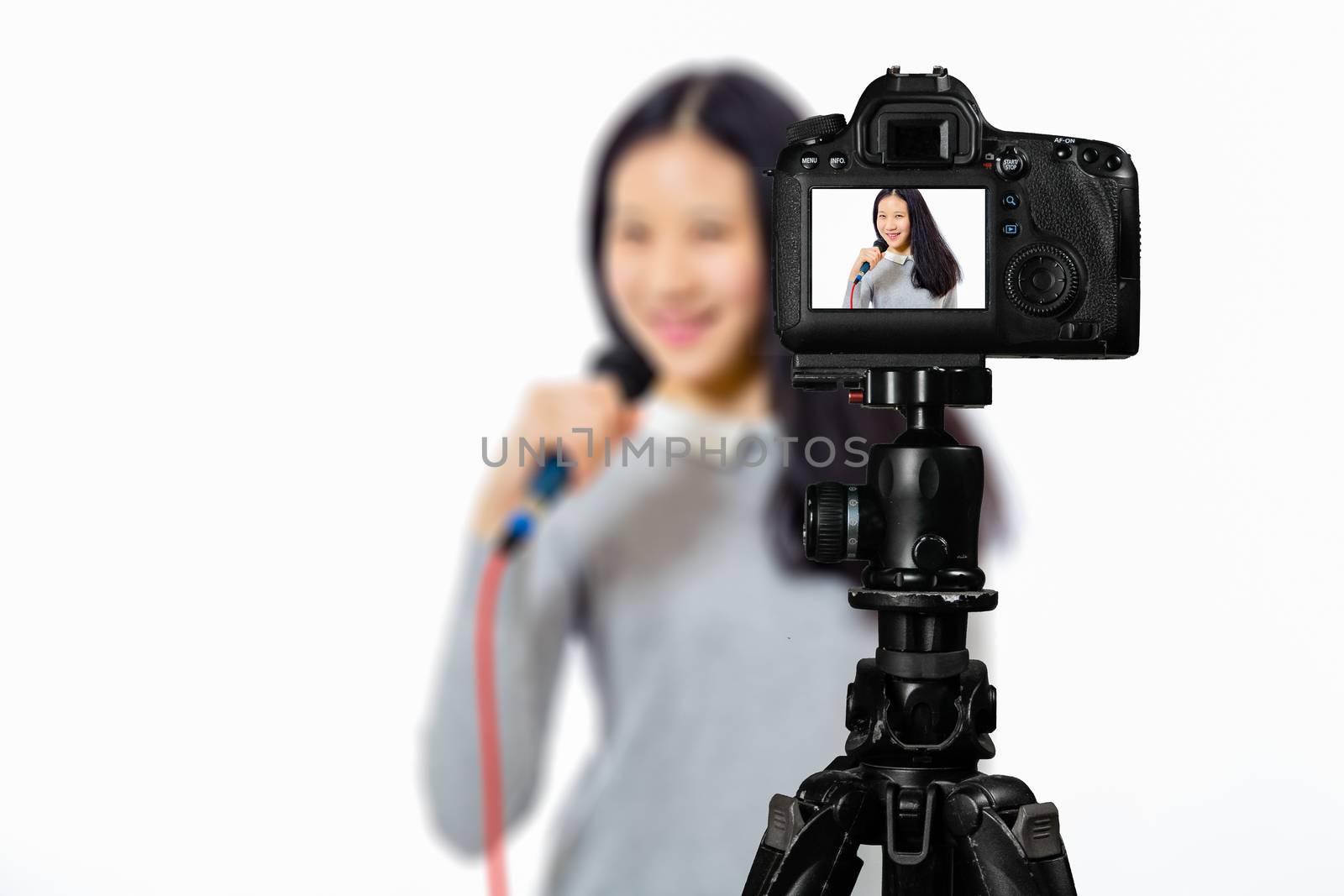 Focus on live view on camera on tripod, teenage girl  singing with microphone image on back screen with blurred scene in background. Teenage vlogger livestreaming show concept
