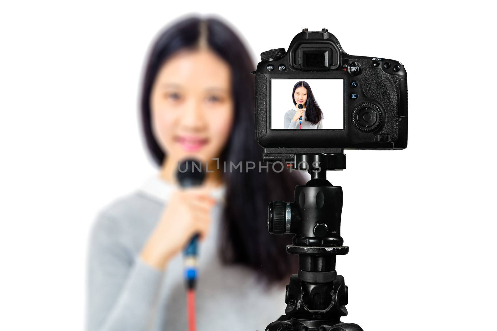 Focus on live view on camera on tripod, teenage girl  singing with microphone image on back screen with blurred scene in background. Teenage vlogger livestreaming show concept