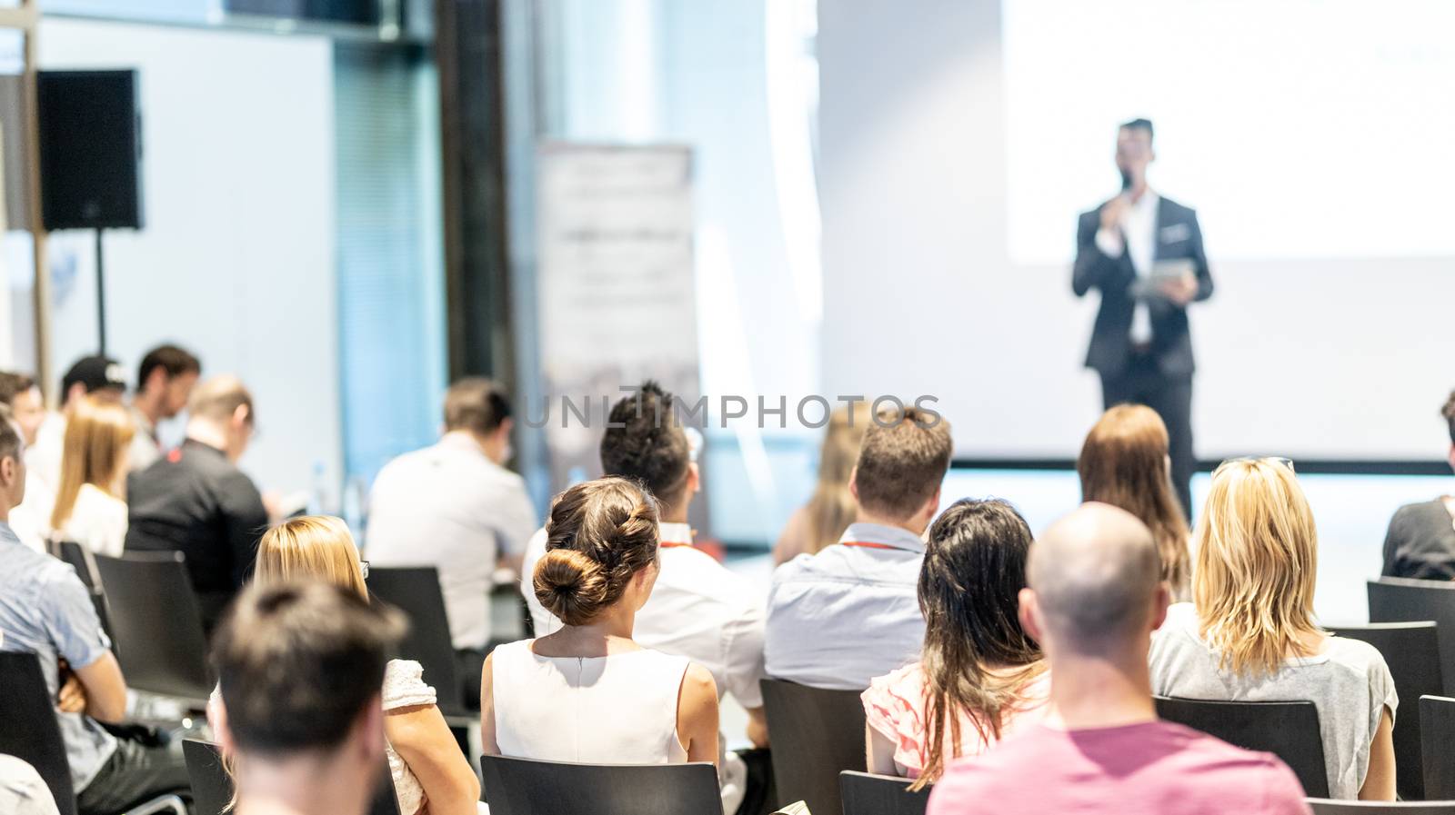 Business and entrepreneurship symposium. Speaker giving a talk at business meeting. Audience in conference hall. Rear view of unrecognized participant in audience.