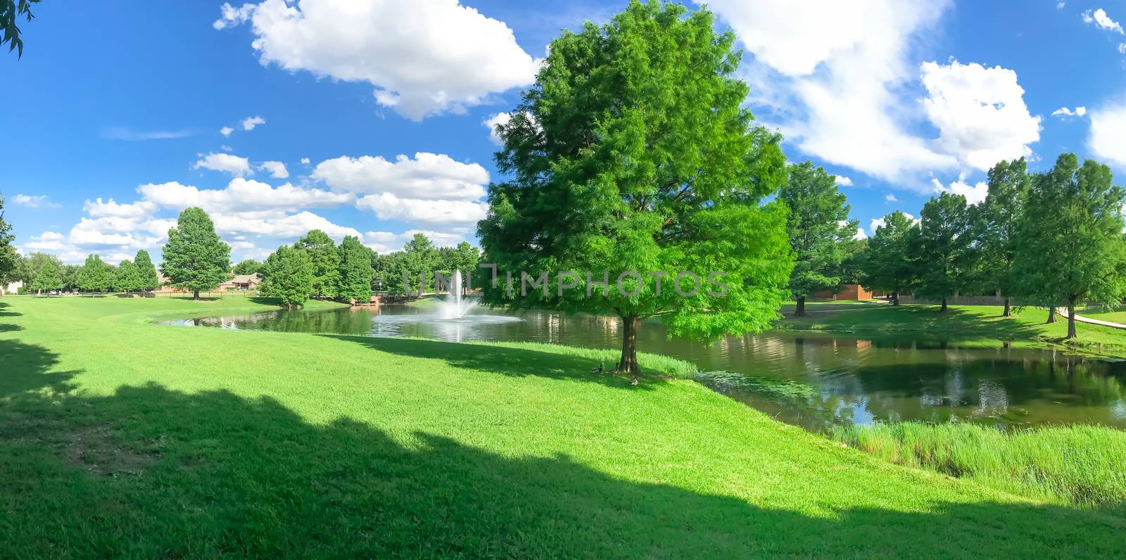 Panorama view beautiful pond with water fountain in small neighborhood North of Dallas, Texas, America. Lake house surrounding by matured trees, green grass lawn and cloud blue sky