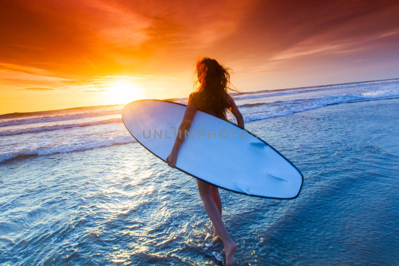 A beautiful young slim sporty woman in bikini with a surfboard is standing at ocean beach at sunset