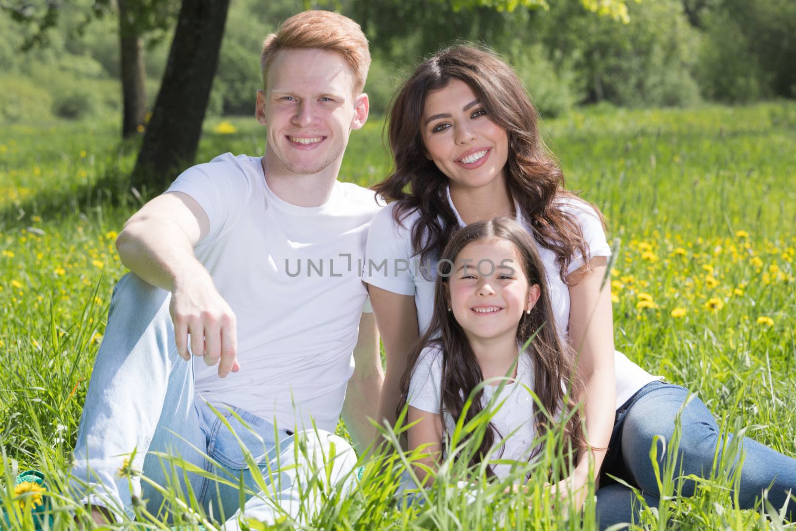 Portrait of happy smiling family of parents and girl sitting on grass with dandelion flowers at sunny summer day