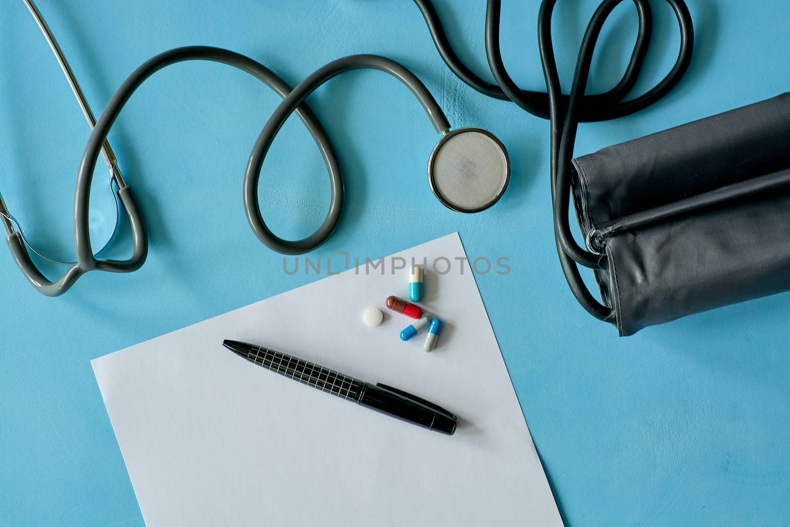 White sheet paper with black pen and colored different pills, capsules, phonendoscope stethoscope, sphygmomanometer on blue background with copy space for text. Medicine consept.