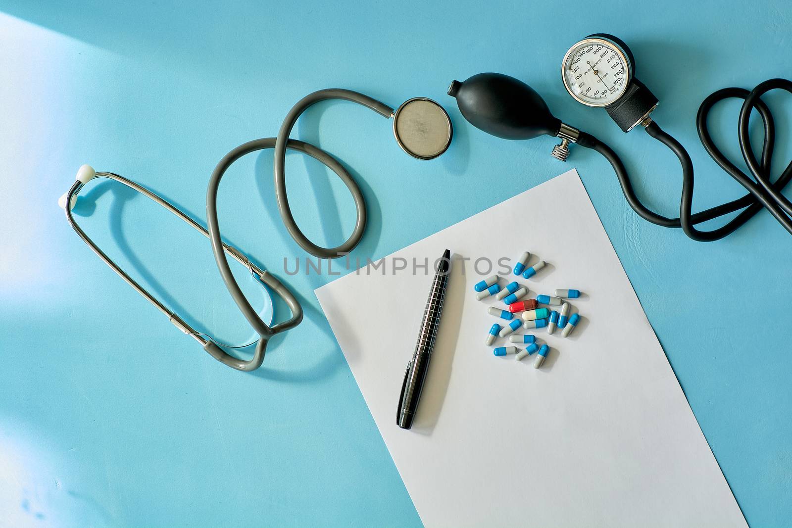 White sheet paper with black pen and colored different pills, capsules, phonendoscope stethoscope, sphygmomanometer on blue background with copy space for text. Medicine consept.