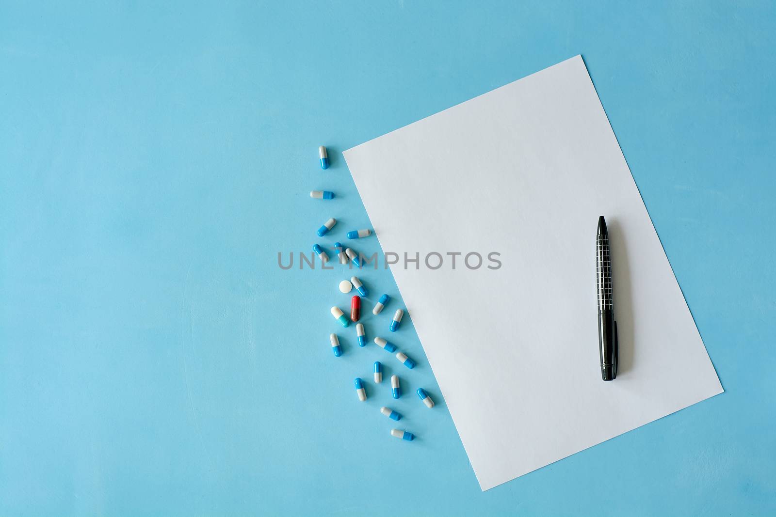 White sheet paper with black pen and colored different pills, capsules on blue background with copy space for text. Medicine consept. Print disign consept.