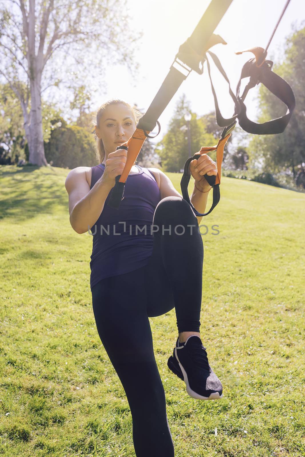 vertical photo of a young beautiful woman doing exercises outdoors under a tree, fitness girl workout with suspension equipment at the morning in a sunny park