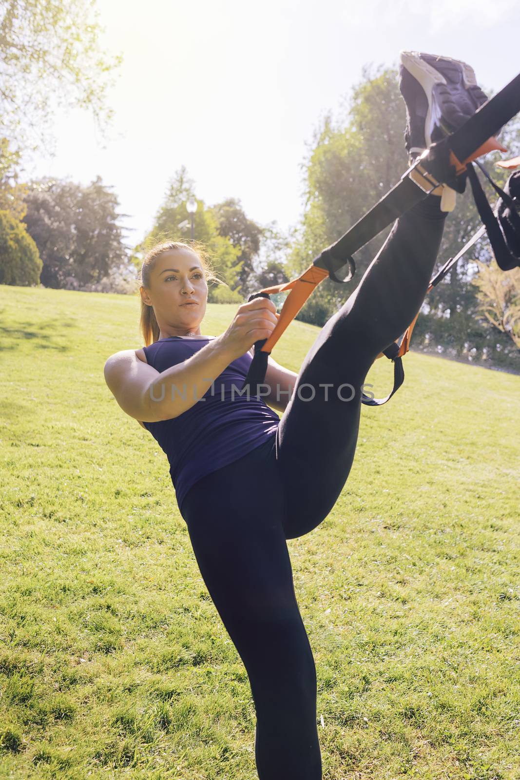 vertical photo of a beautiful young woman doing exercises outdoors under a tree, fitness girl workout with suspension straps at the morning in a sunny park