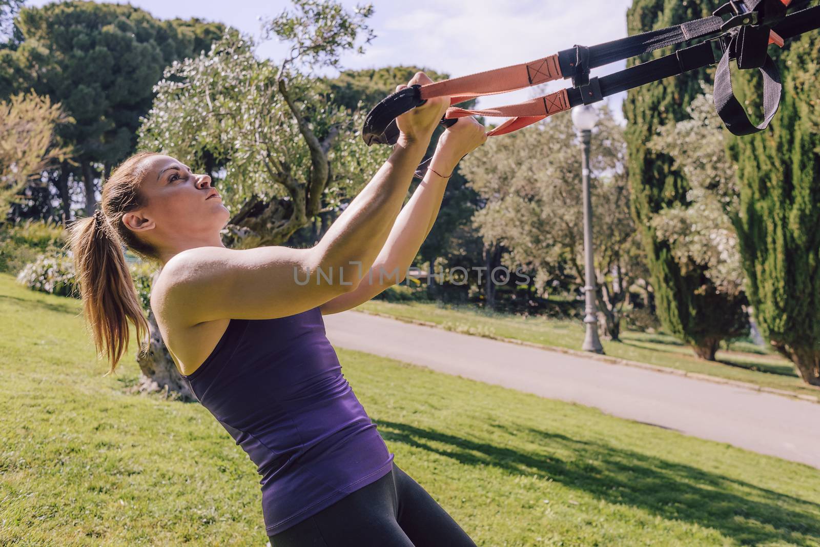 athletic young woman doing bicep curls with fitness suspension straps, during functional workout in a outdoor park
