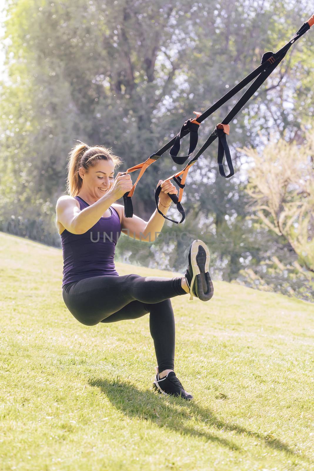 vertical photo of a strong young woman that smiles training with fitness suspension straps, during functional workout in a sunny outdoor park at the morning