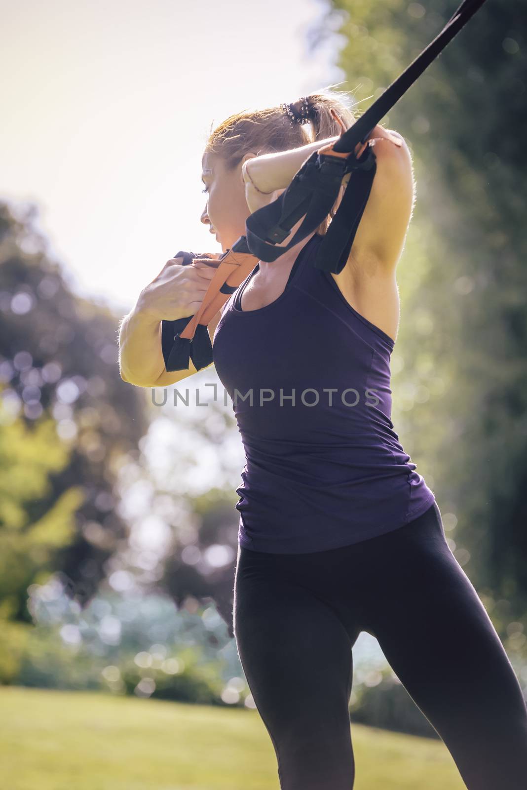 vertical photo of an athletic woman training with fitness straps, during functional workout in a sunny outdoor park in the morning