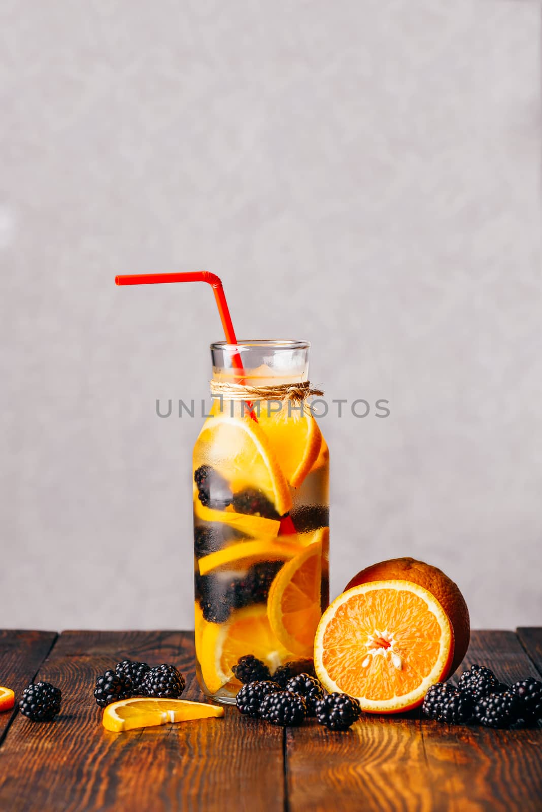 Red Straw in Bottle of Chilled Water Infused with Sliced Raw Orange and Fresh Blackberry. Ingredients on Wooden Table.