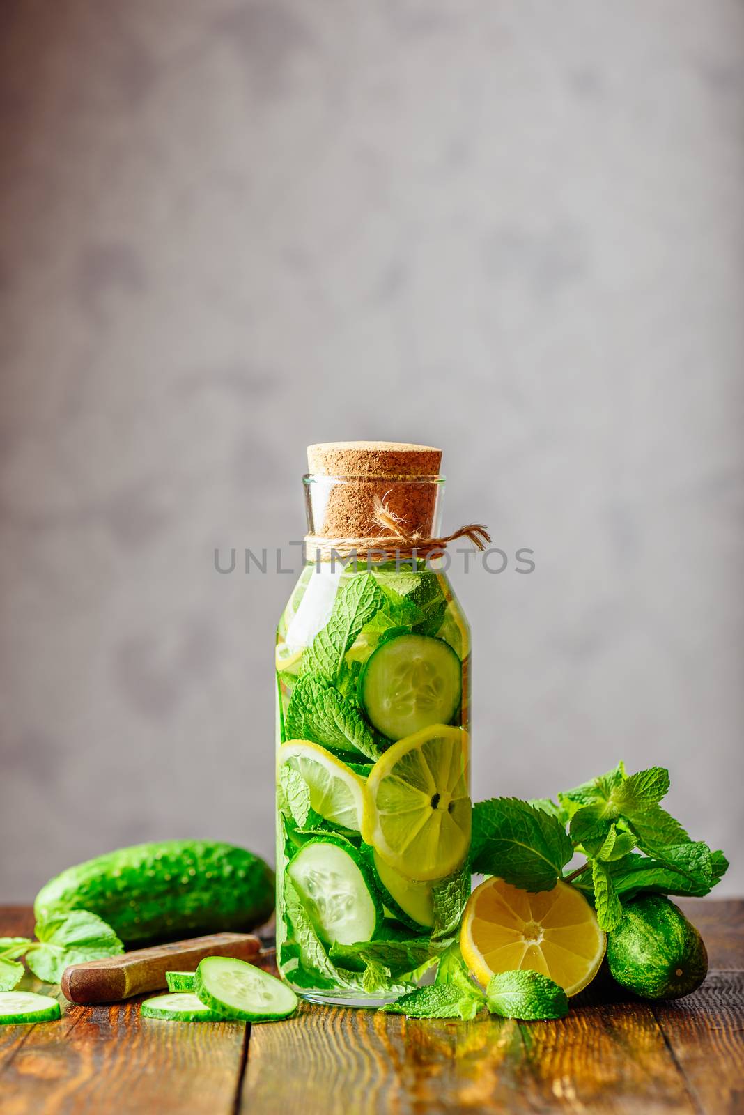 Bottle of Water Infused with Sliced Lemon, Cucumber and Mint Leaves. Ingredients and Knife on Table. Copy Space on the Top. Vertical Orientation.