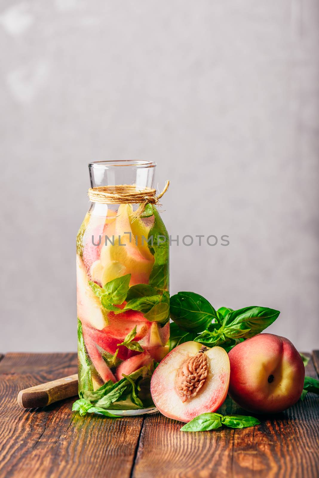 Bottle of Infused Water with Sliced Peach and Basil Leaves. Knife and Ingredients on Wooden Table. Vertical Orientation.