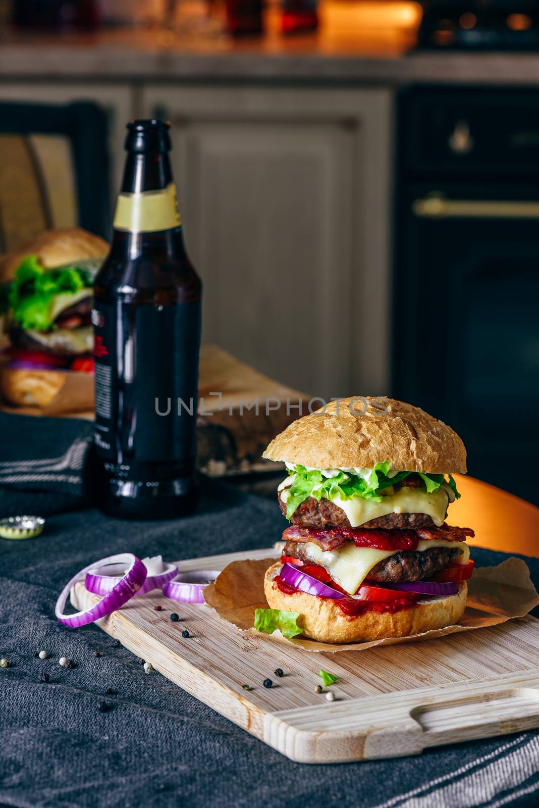 Prepared Cheeseburger with Two Beef Patties on Cutting Board. Bottle of Beer and Some Ingredients on Table.