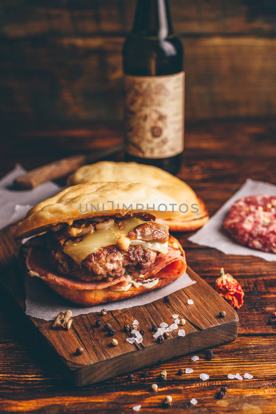 Homemade Burger on Cutting Board. Cheeseburger with Beef Patty, Wisconsin Swiss Cheese, Ham, Sauteed Mushrooms, Dijon Mustard, Mayonnaise and Potato Roll. Bottle of Craft Beer on Background.