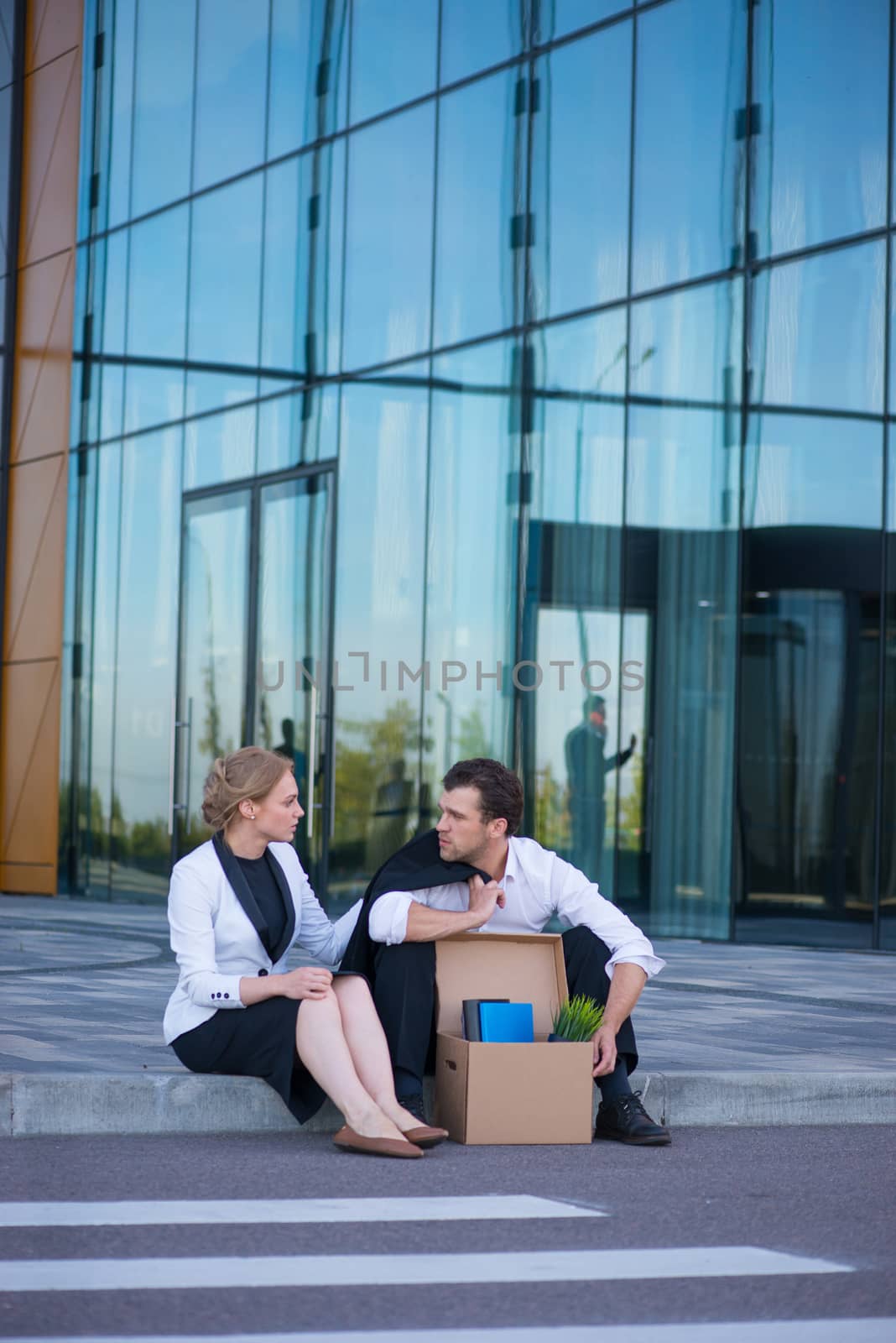 Fired business man sitting frustrated and upset on the street near office building with box of his belongings. He lost work. Business woman comforts and encourages him