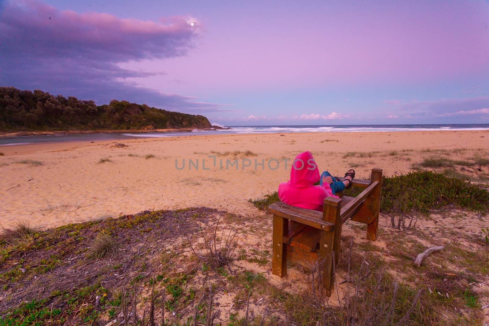 Woman relaxes on timber bench overlooking beach in the cool hues of dusk