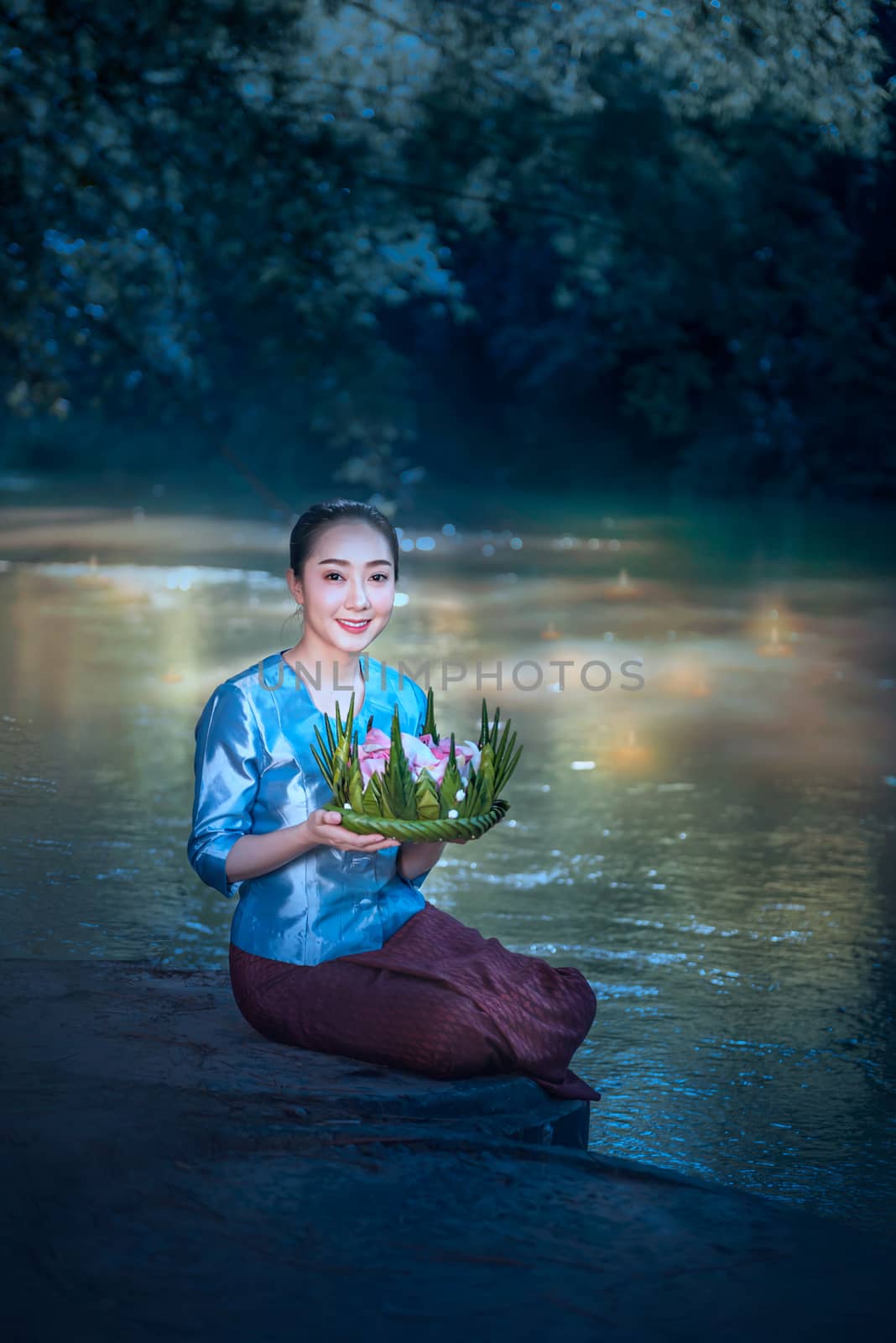 beautiful woman in traditional asian dresses holding vessel made of banana leaves and flowers sitting near river ( Loy Krathong Traditional Festival )