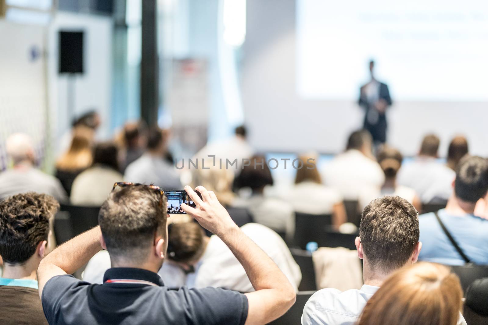 Business and entrepreneurship symposium. Speaker giving a talk at business meeting. Audience in conference hall. Rear view of unrecognized participant in audience taking photo of presentation.