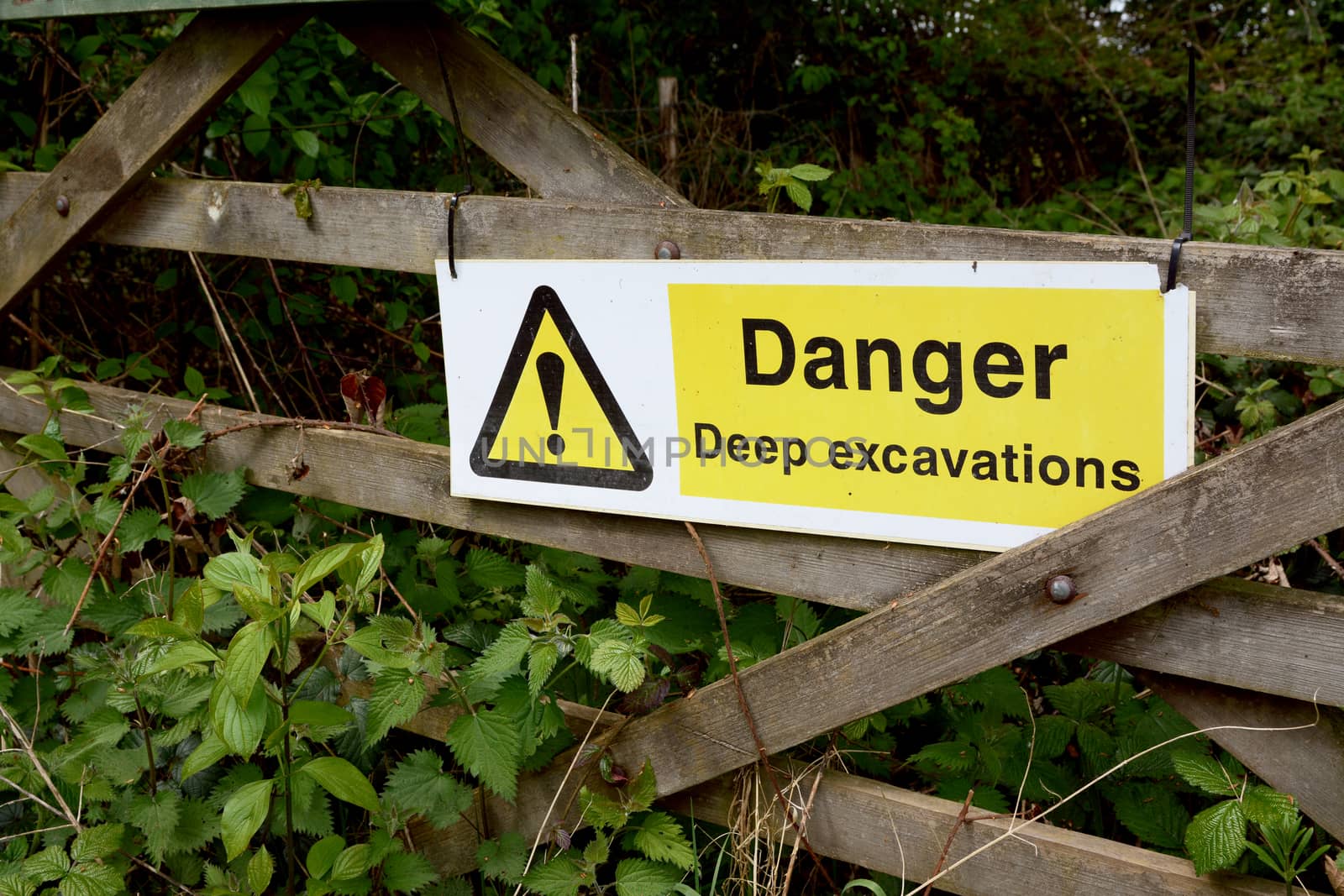 Warning sign - Danger Deep Excavations - fixed to a rustic wooden gate, overgrown with nettles in the countryside