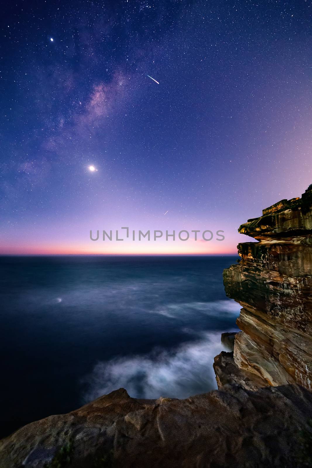 Sydney coast by night with starry universe and milky way overhead and a deep blue ocean p9unding the sandstone cliffs of Sydney East Coast of Australia