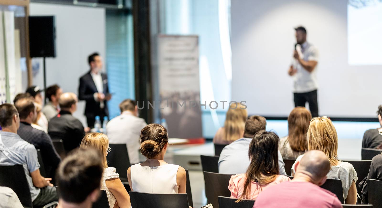 Business and entrepreneurship symposium. Speaker giving a talk at business meeting. Audience in conference hall. Rear view of unrecognized participant in audience.