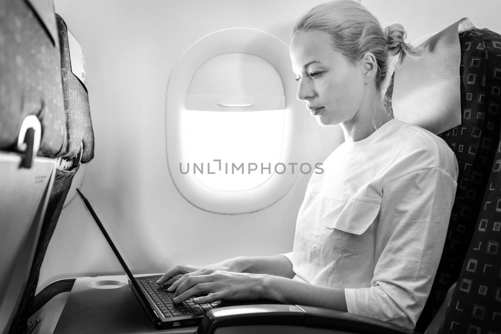 Attractive caucasian female passenger working at modern laptop computer using wireless connection on board of commercial airplane flight. Black and white image.