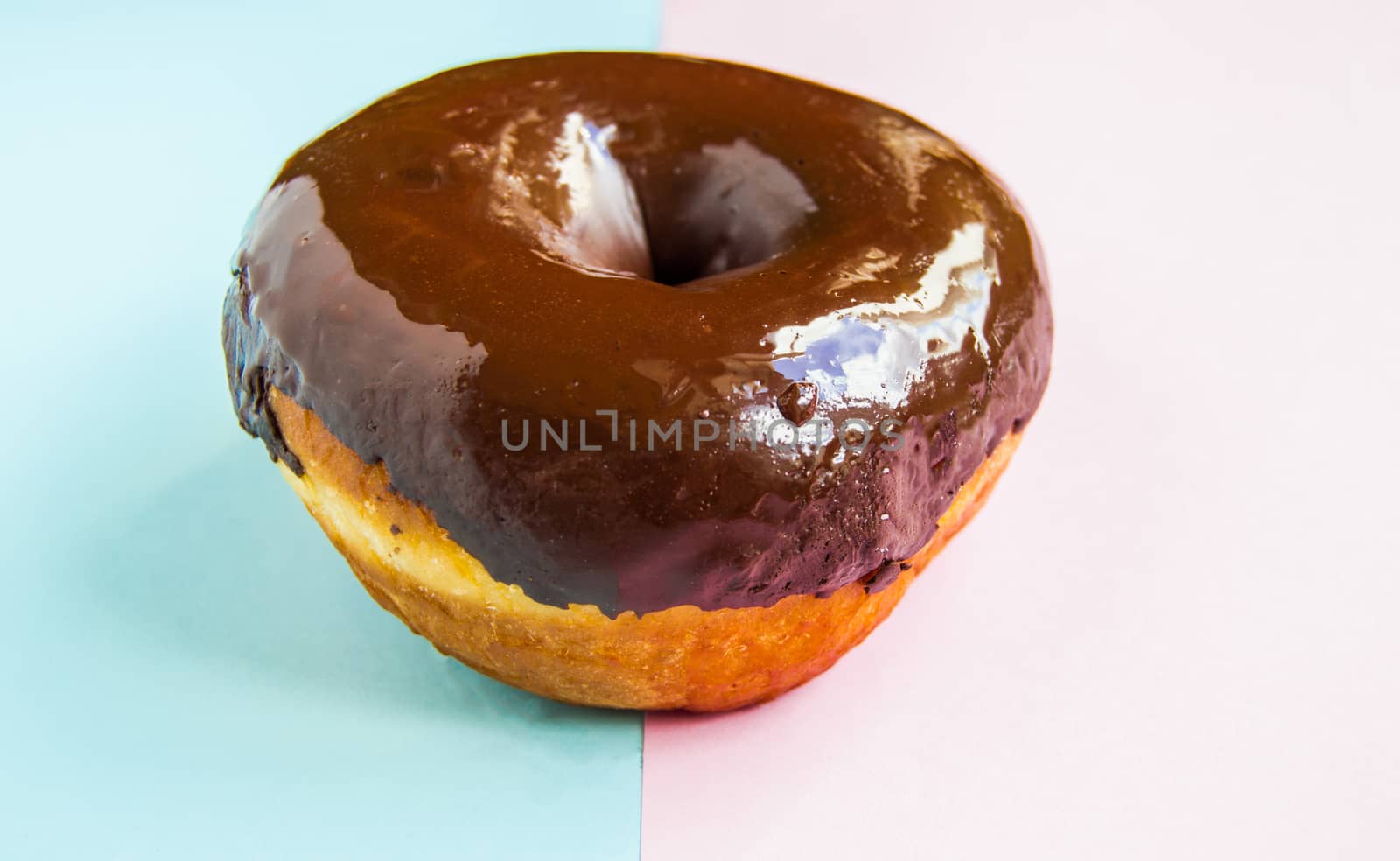 Delicious donut with shiny chocolate icing on pastel pink-blue background, close-up, side view.
