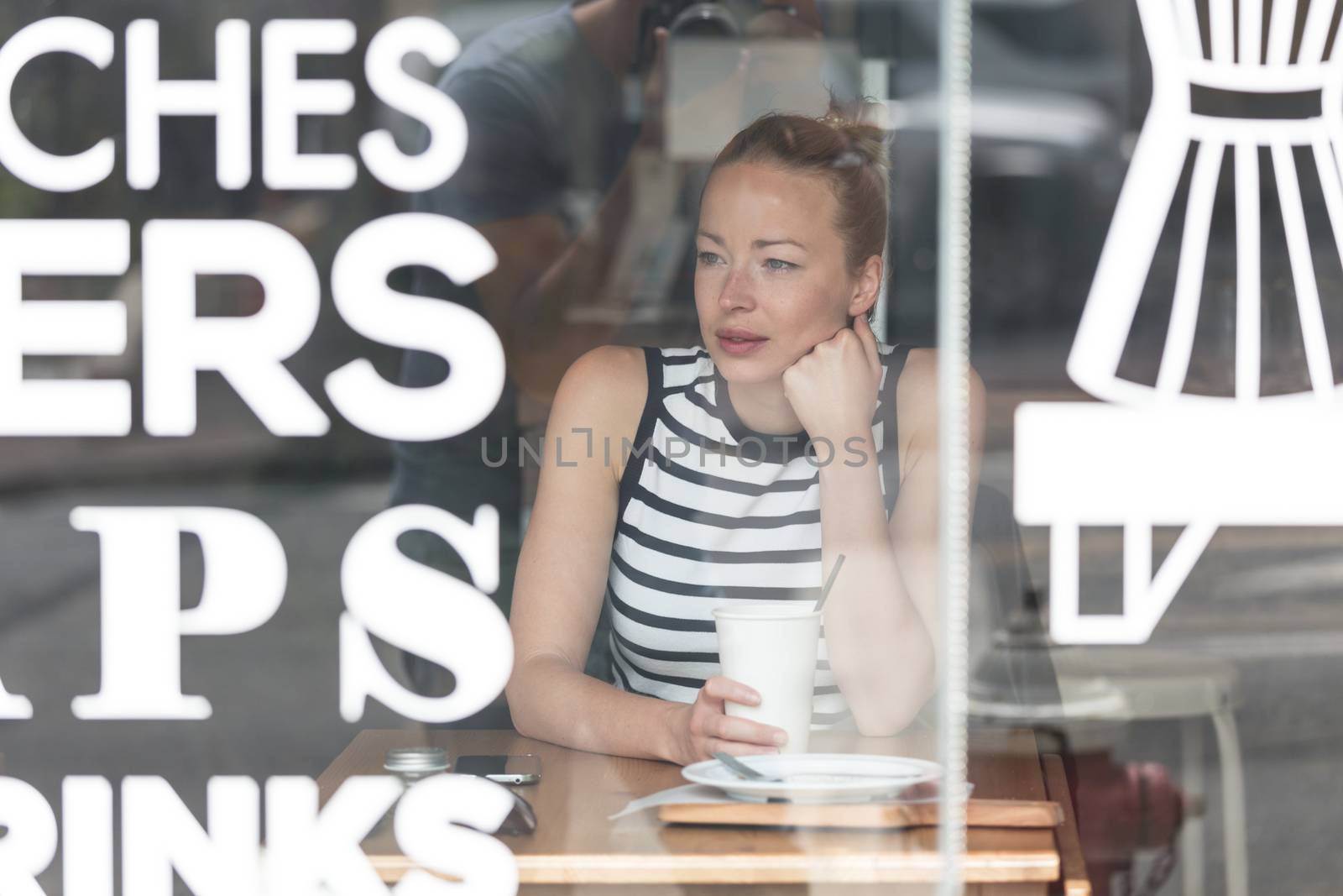 Young caucasian woman sitting alone in coffee shop thoughtfully leaning on her hand, looking trough the window by kasto