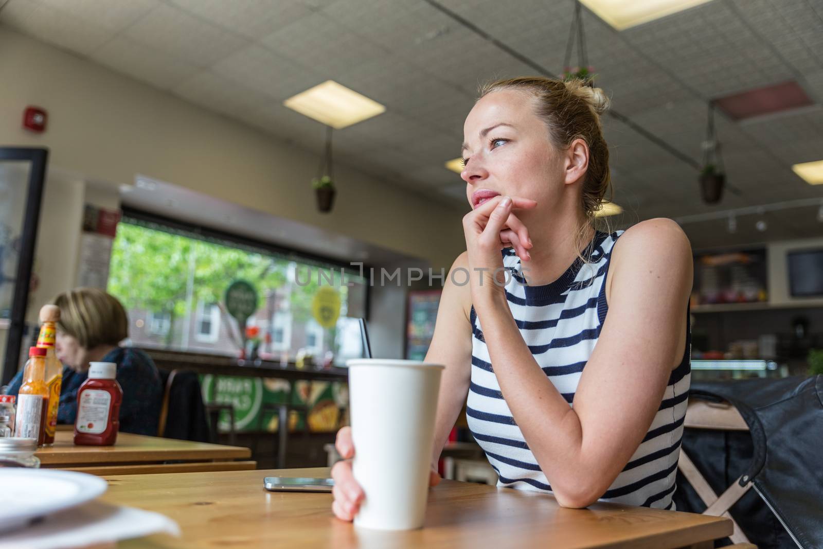 Young caucasian woman sitting alone in coffee shop thoughtfully leaning on her hand, looking trough the window by kasto