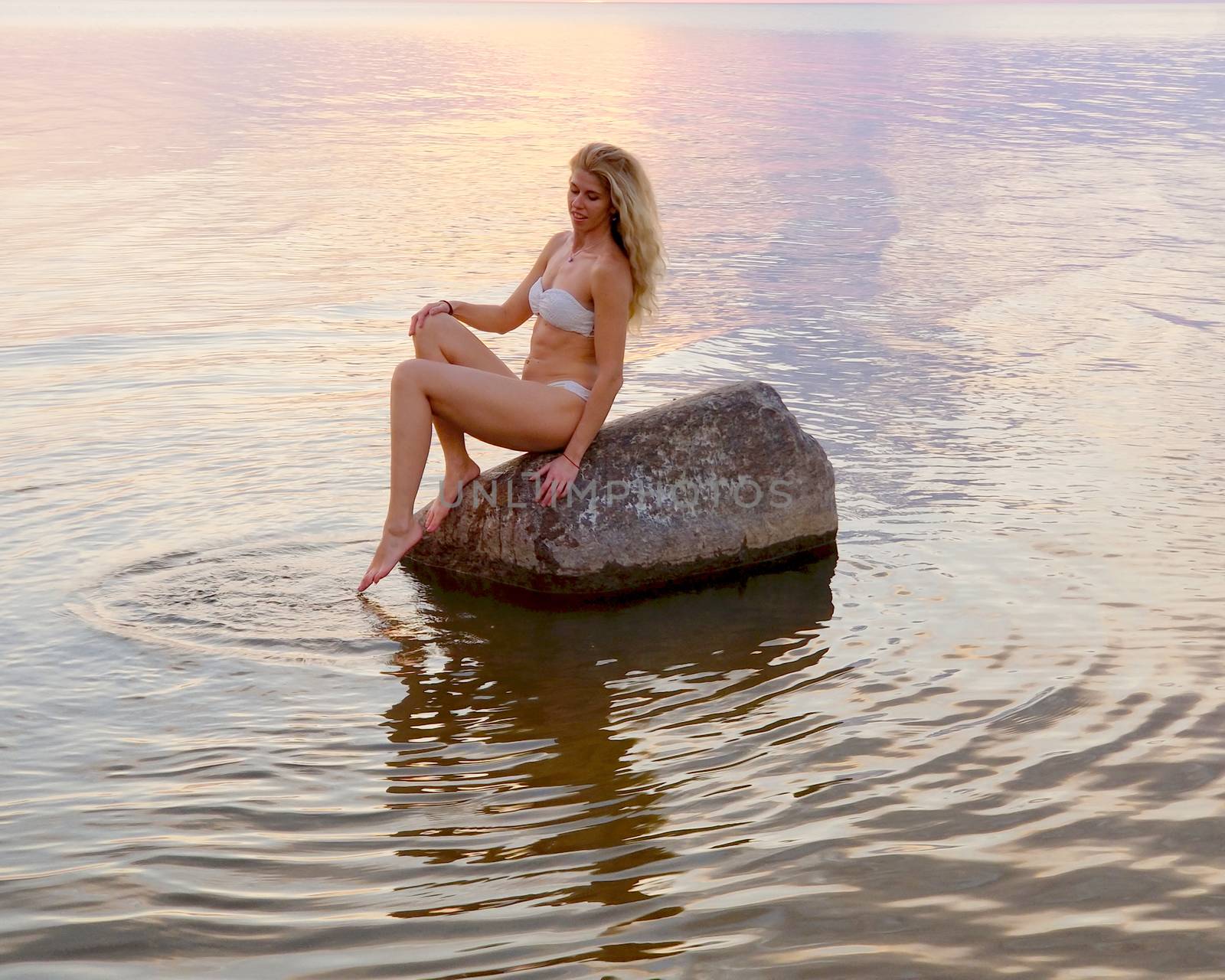 beautiful tanned blonde in a white bikini sits on a stone standing in the water during sunset.