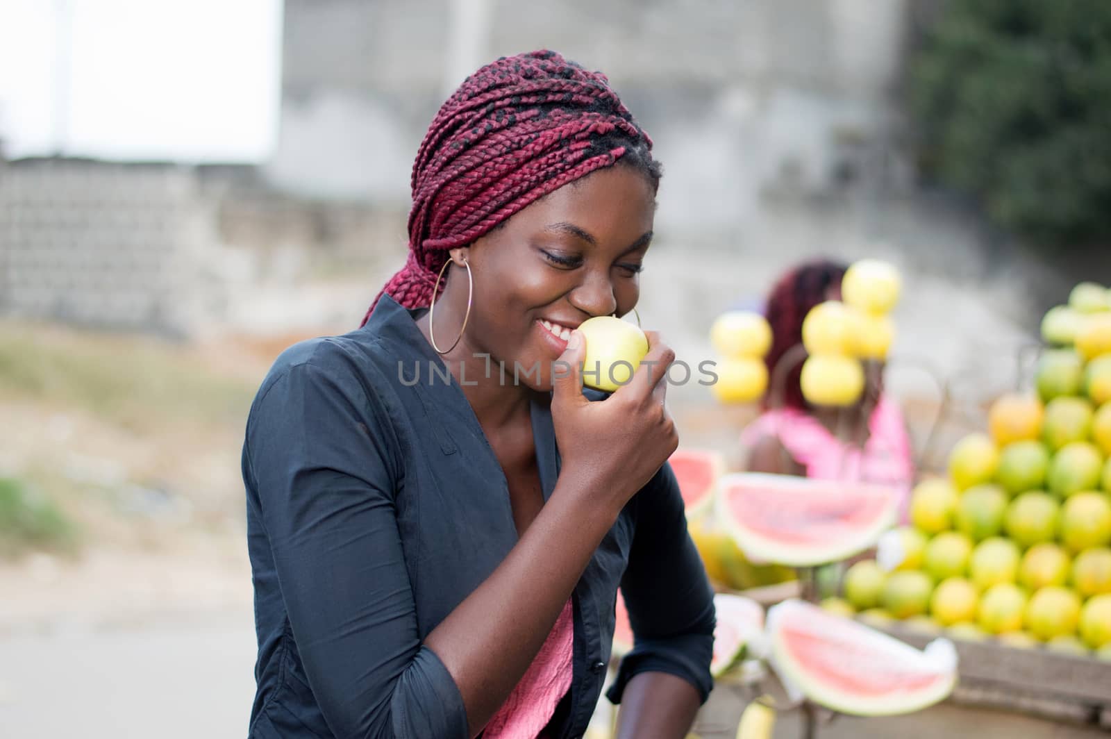 smiling young woman standing near a fruit shelf is eating an apple.
