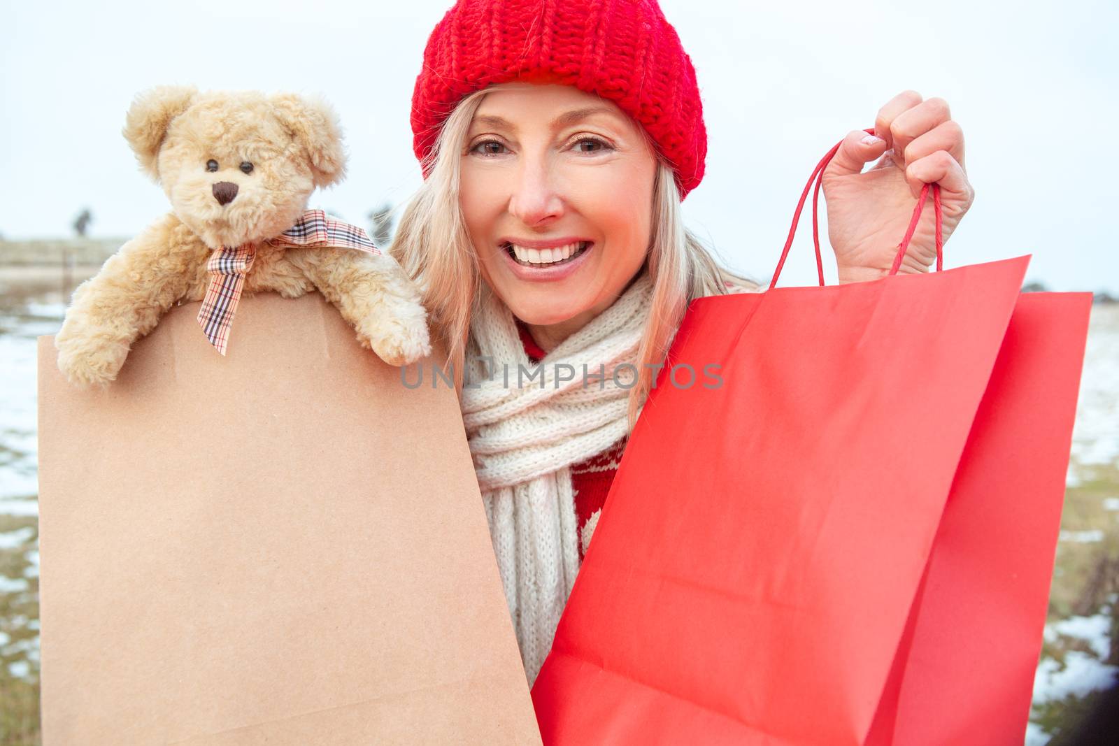 Winter woman holding shopping bags filled with goodies. A generic brown bear poking out of one. She is wearing winter clothing with a knitted beanie and scarf in a winter landscape.