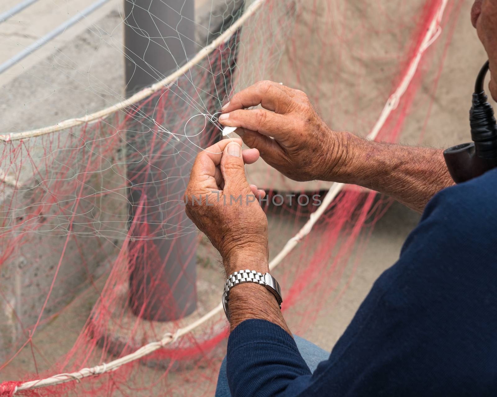 Old fisherman reparing fishing net, sitting with pipe in his mouth.