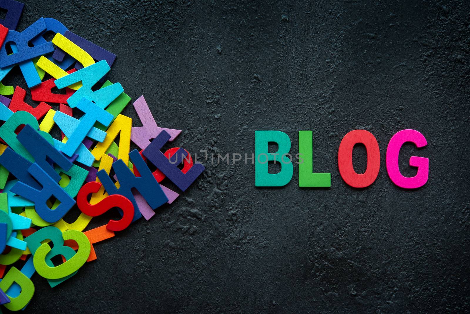 The colorful words "BLOG" made with wooden letters next to a pile of other letters over dark background.